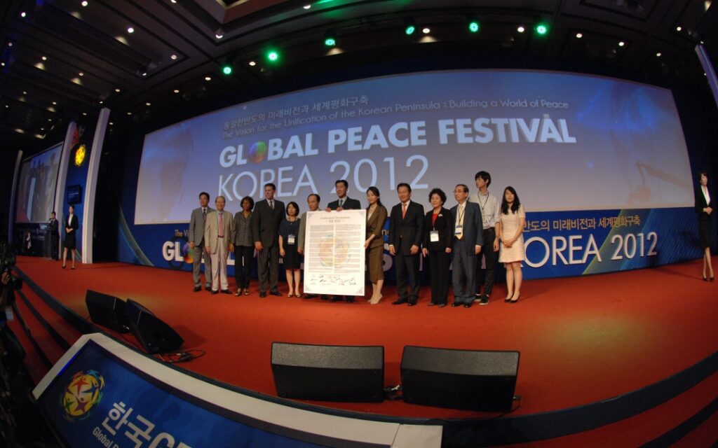 A group of people stands on stage holding a large document at the Global Peace Festival Korea 2012, with event banners in the background.
