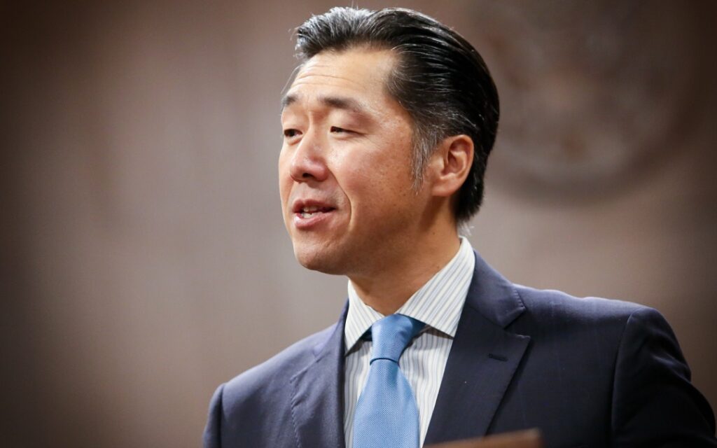 A man in a suit and blue tie speaking in front of a blurred brown background.