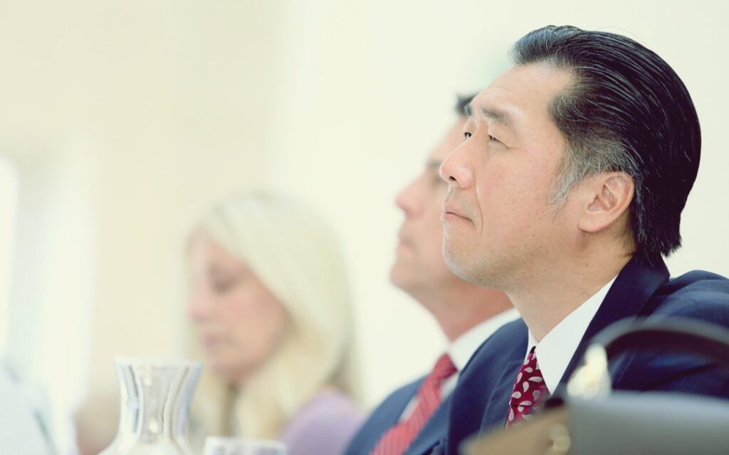 A man in a suit sits attentively in a meeting at the Action for Korea United Fifth Anniversary Assembly, with two other blurred people and a glass pitcher visible in the background.