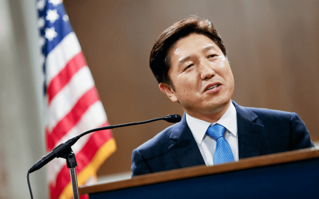 A man in a suit stands at a podium with a microphone, speaking about innovative approaches, with a blurred U.S. flag in the background.