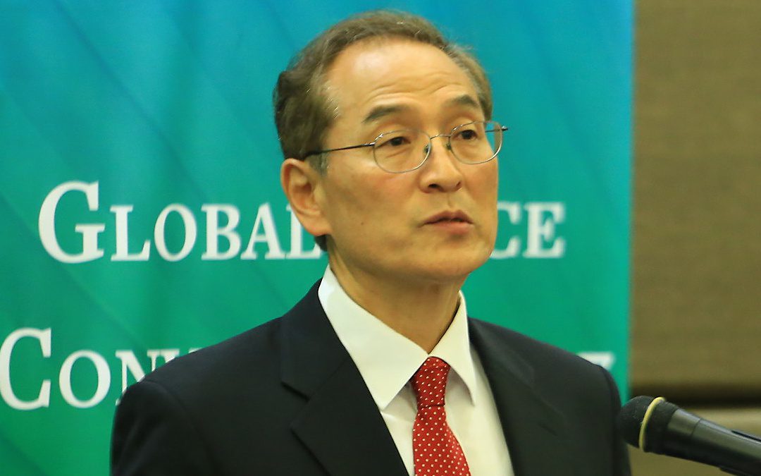 A man in a suit and red tie speaks at a podium in front of a blue-green background with the partially visible words