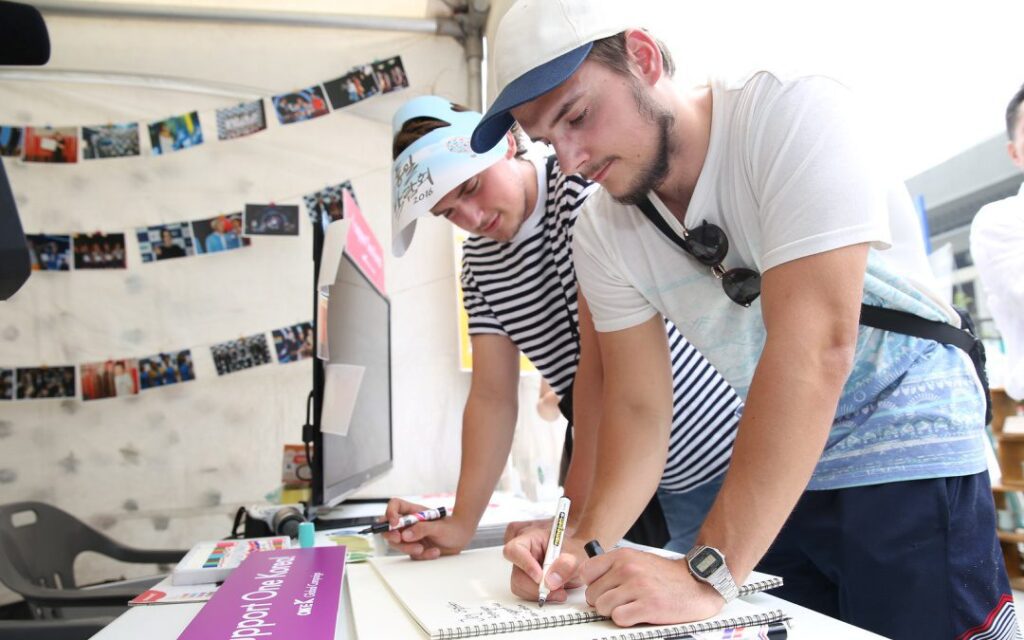 Two young men write with markers on paper at an outdoor booth, highlighting civil society's emerging role, with photos hanging on a string in the background.