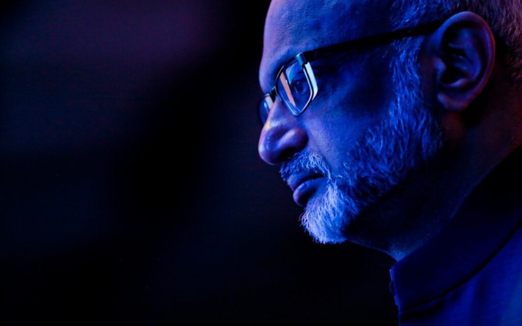 A man with glasses and a beard is pictured in profile under dramatic blue lighting, looking thoughtful.