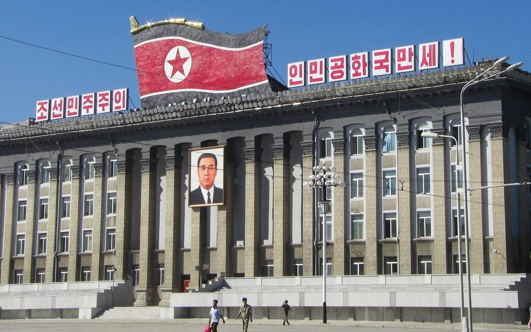 A large building with columns displays a North Korean flag, Korean banners, and a portrait of a man above the entrance; people walk in front.