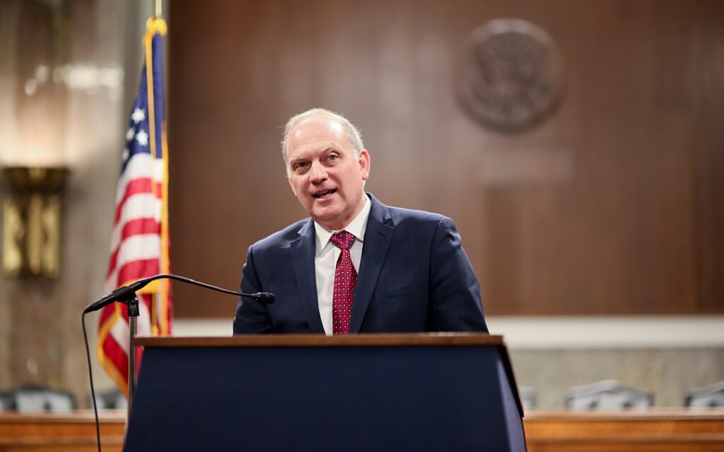 A man in a suit speaks at a podium in a formal setting, with an American flag and an official seal visible in the background.
