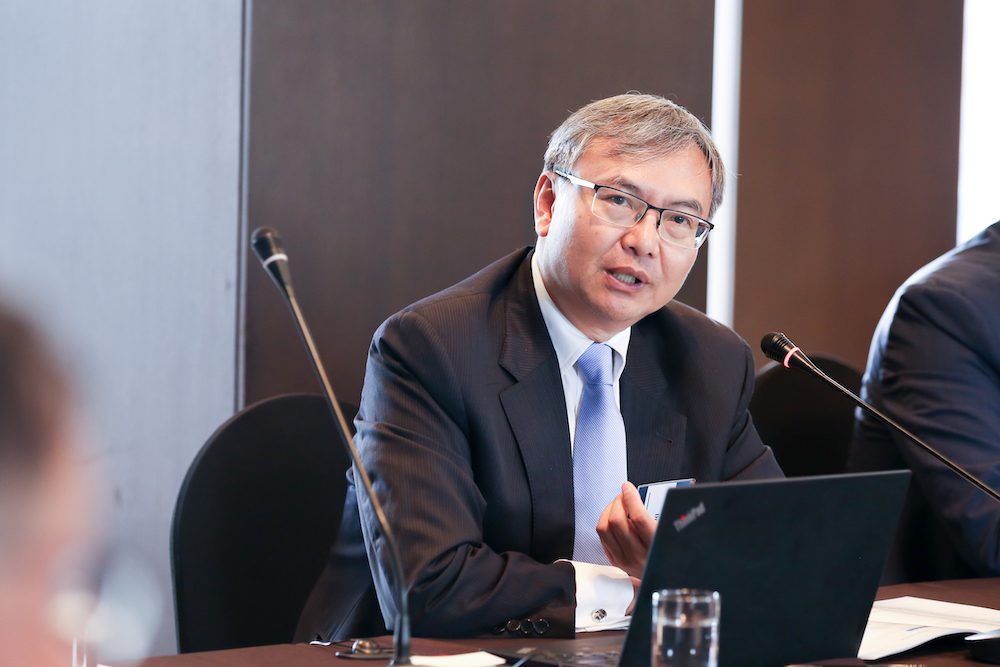 A man in a suit and tie speaks into a microphone at a meeting table, discussing policy on denuclearization in Northeast Asia, with a laptop and a glass of water in front of him.