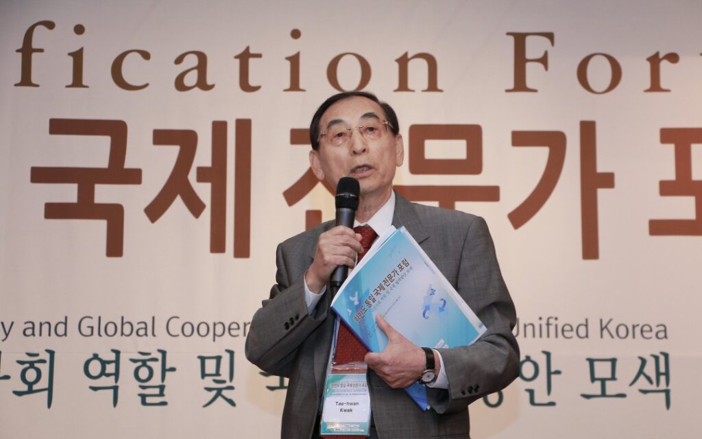 An elderly man in a suit holds a microphone and a blue booklet, speaking on stage in front of a banner with Korean and English text.