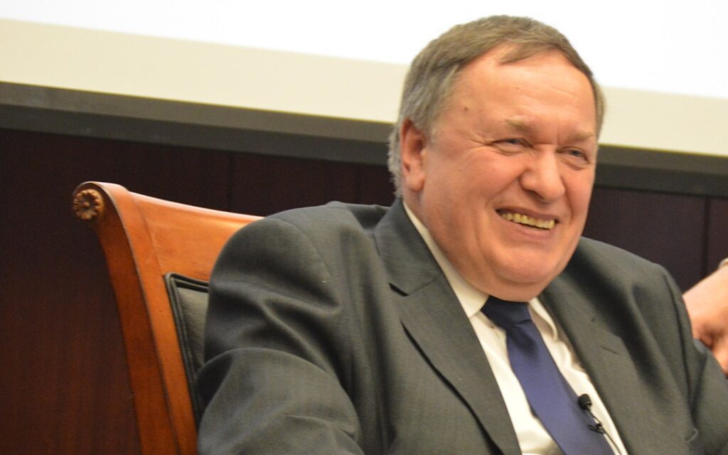 A man in a suit and tie sits in a wooden chair, smiling and looking to the side during an event or discussion.