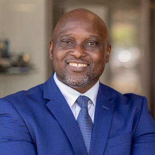 A man in a blue suit and tie stands indoors, smiling with his arms crossed.