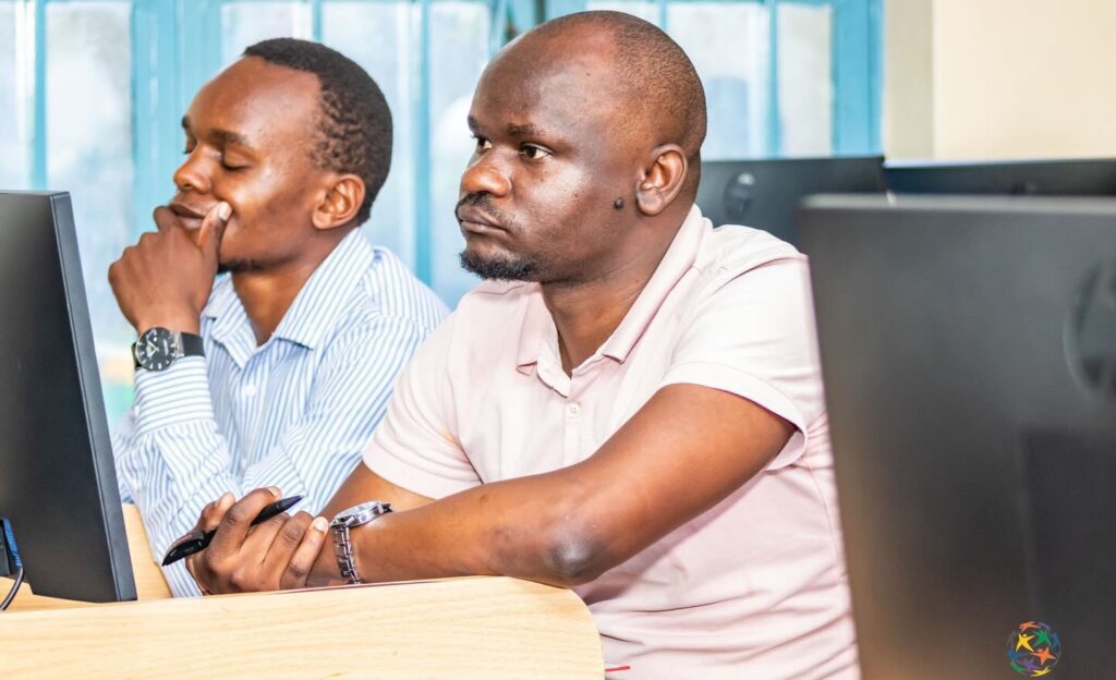 Two men sit at computer desks in a classroom—one, likely among Computer Science Educators, appears deep in thought with his hand on his chin, while the other looks attentively forward holding a pen.