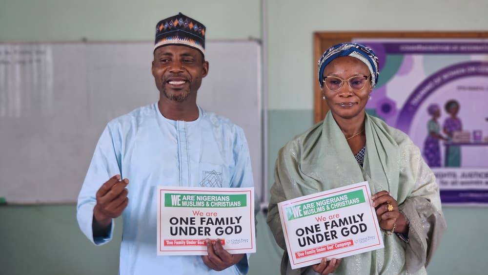 Two people stand indoors in Kaduna holding signs that read “We are Nigerians, Muslims & Christians. We are one family under God,” celebrating unity for International Women’s Day 2026 with GPF Nigeria.