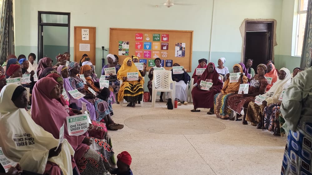 A group of women sit in a large circle indoors in Kaduna, many holding signs, with posters displayed on the wall behind them—gathered for GPF Nigeria’s International Women’s Day 2026 event.
