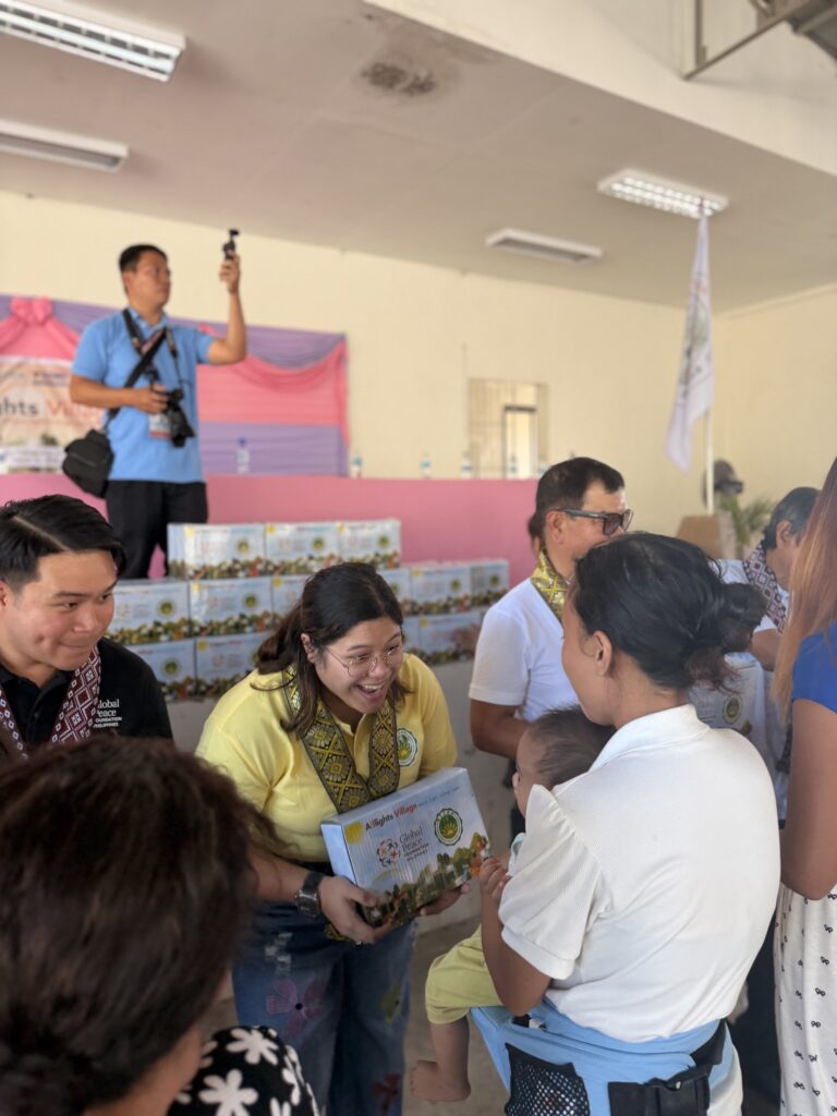 In Batangas, a group from GPF Philippines and All-Lights Village, some wearing sashes, distribute “Manna Pack” boxes to a woman holding a child in an indoor setting with more boxes stacked in the background.