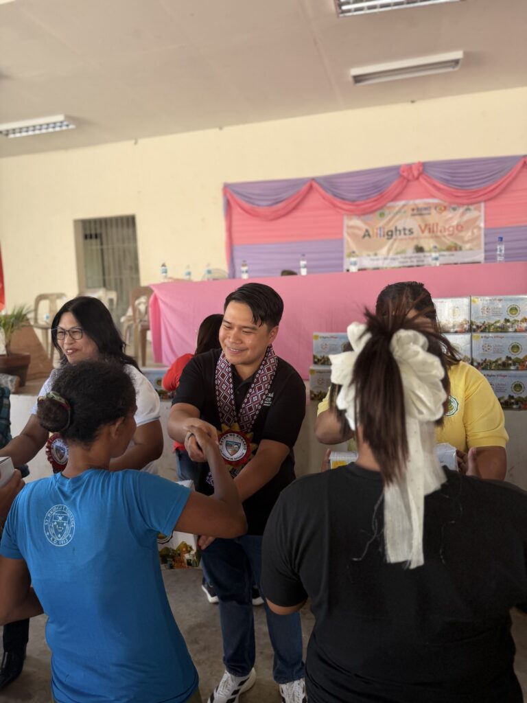 A group of people stands indoors in Batangas as a man with a garland and medallion faces women, handing out packages at an All-Lights Village distribution event organized by GPF Philippines.