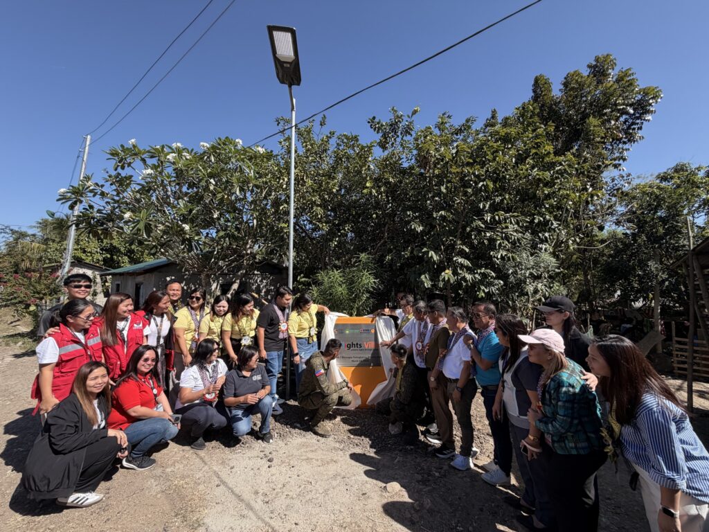 A group of people poses outdoors near a lamp post and a yellow sign in Batangas, with trees and a building in the background under a clear blue sky, representing the spirit of All-Lights Village and GPF Philippines.