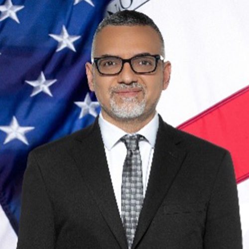 A man in a suit and tie stands in front of overlapping United States and California state flags.