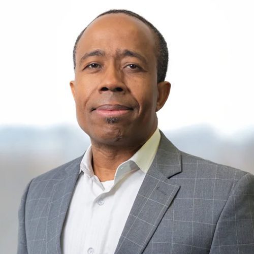 A man wearing a gray checked suit jacket and white shirt poses against a blurred background.