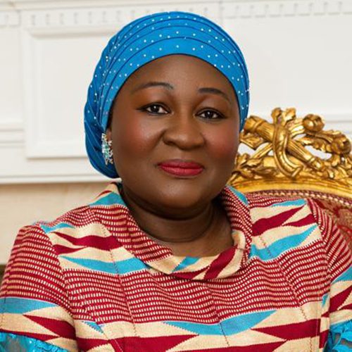 A woman wearing a blue patterned headscarf and a colorful patterned dress sits on an ornate gold chair, smiling at the camera.