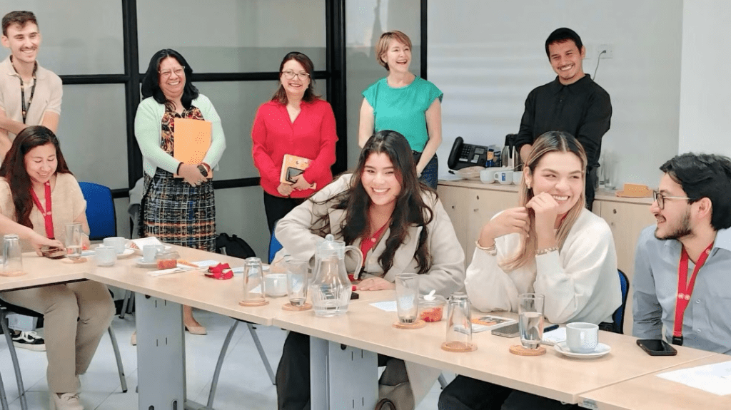 A group of people, some seated at a conference table with snacks and drinks, and others standing, smiling and engaging in conversation about Youth Digital Leadership in a meeting room in Guatemala.