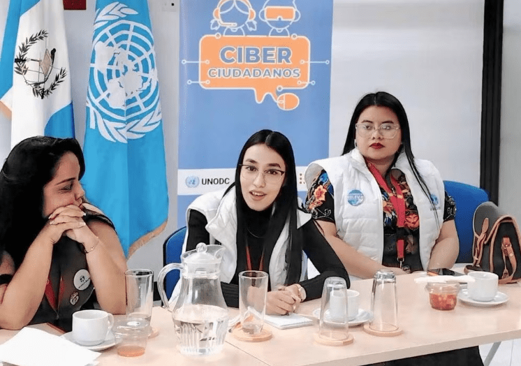 Three women sit at a conference table with drinks and snacks, in front of a