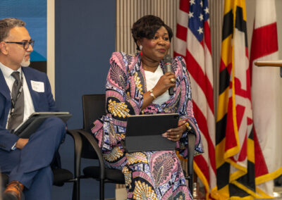 Two people sit on a panel; the woman in colorful attire holds a microphone and tablet, while the man in a suit holds a tablet. US and Maryland flags are in the background at the Ubuntu Africa Diaspora Summit 2026.