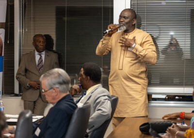 A man in a tan outfit speaks into a microphone while standing in a conference room at the Diaspora Summit; other people are seated and listening attentively.