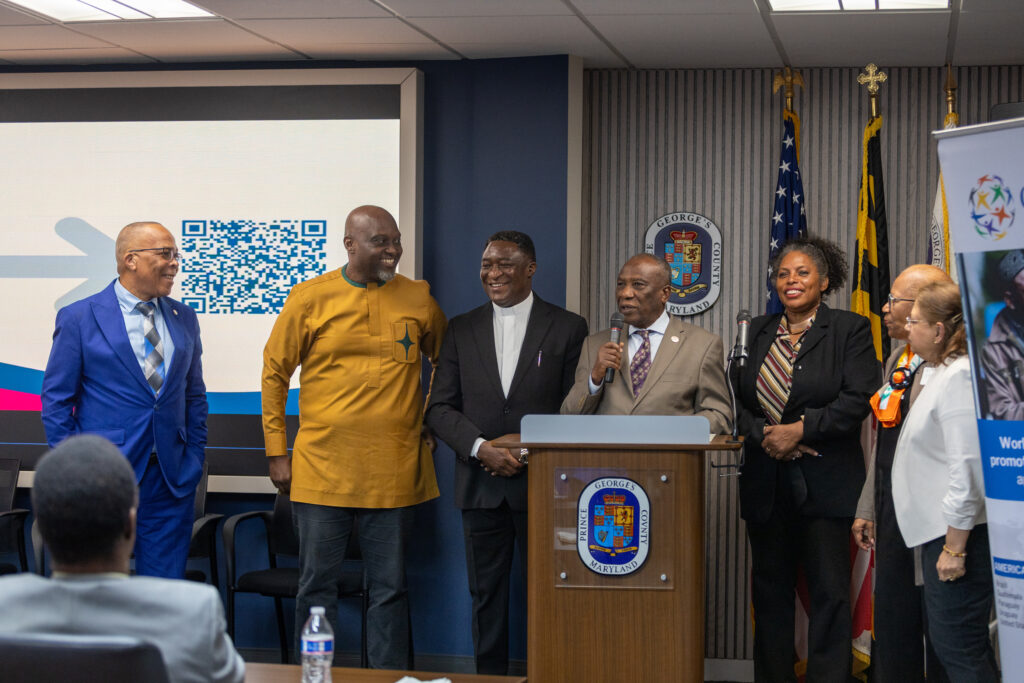 A group of people in formal attire stand at a podium during an indoor event, with a QR code displayed on a screen in the background at the Ubuntu Africa Diaspora Summit.