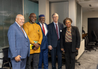 Five professionally dressed people stand together and smile for a group photo in an office setting with large windows and chairs, capturing a moment at the 2026 Ubuntu Africa Diaspora Summit gallery.