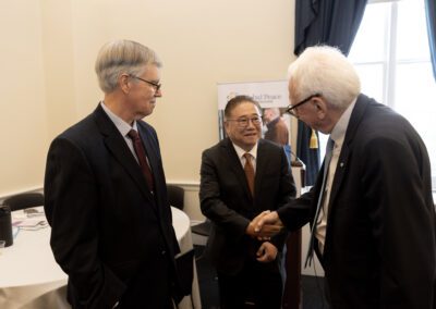 Three men in suits converse in the Capitol Forum Policy Gallery; two are shaking hands while the third stands nearby, smiling.