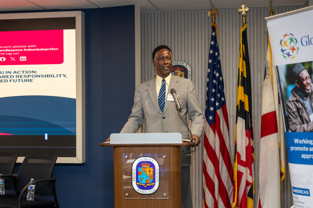 A man in a gray suit speaks at a podium during the 2026 Ubuntu Africa Diaspora Summit, with multiple flags and a presentation screen behind him at the indoor gallery event.