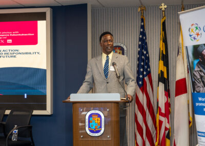 A man in a gray suit speaks at a podium during the 2026 Ubuntu Africa Diaspora Summit, with multiple flags and a presentation screen behind him at the indoor gallery event.