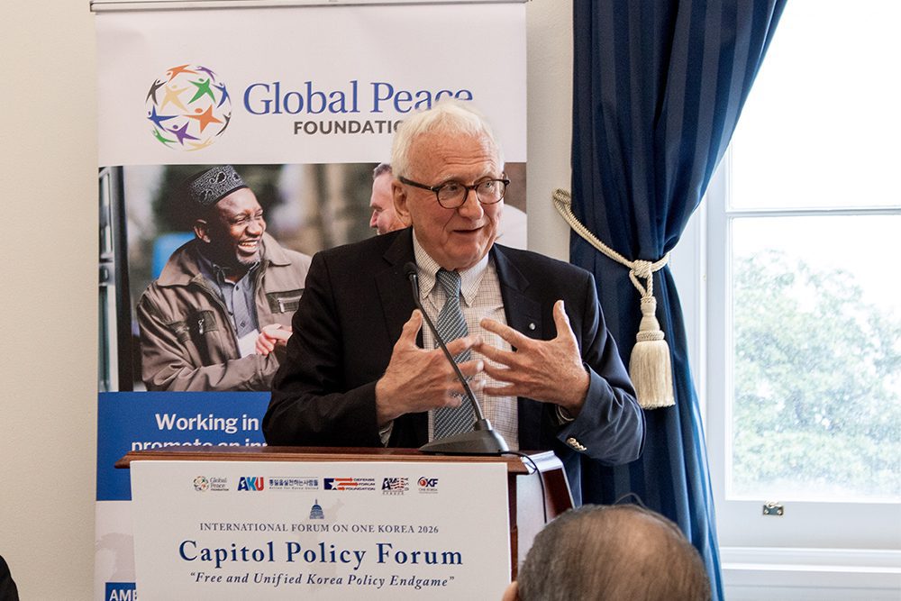 An older man speaks at a podium during the Capitol Policy Forum, with a Global Peace Foundation banner and an audience member visible.