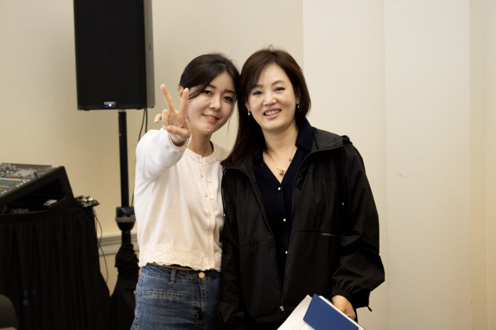 Two women stand indoors at the Capitol Forum Policy Gallery; one smiles holding documents, while the other flashes a peace sign at the camera. Audio equipment is visible in the background.