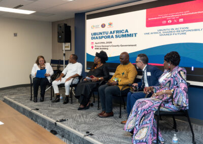 Six panelists sit on stage discussing at the Ubuntu Africa Diaspora Summit, with a screen behind them displaying event details and the Ubuntu Africa Gallery.