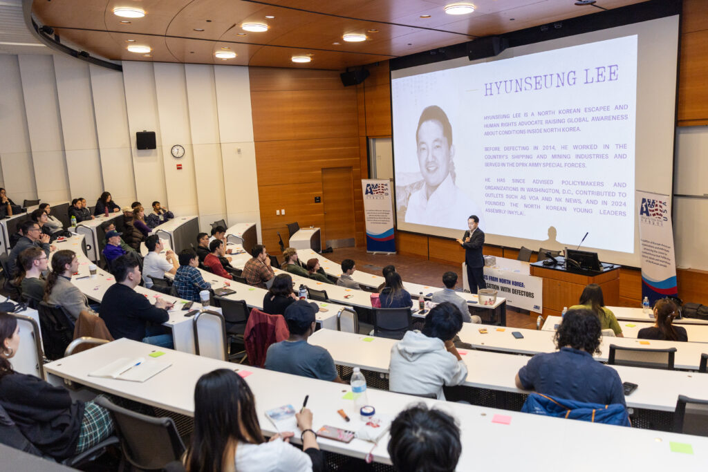 A speaker presents to an audience in a lecture hall at the University of Washington, with a large screen displaying information about Hyunseung Lee, a North Korean escapee and human rights activist, as part of the Voices from North Korea series.