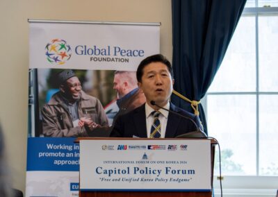 A man in a suit speaks at a podium during the Capitol Policy Forum, connecting themes of the International Forum on One Korea, with a Global Peace Foundation banner and window in the background.