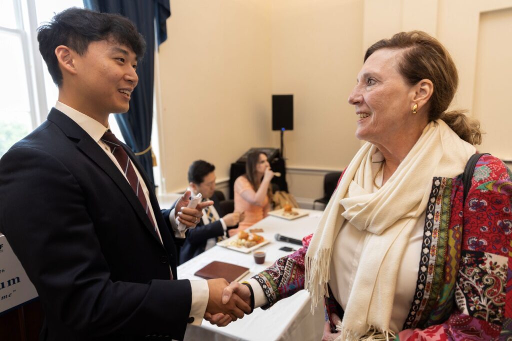 A man in a suit and a woman in a colorful jacket and scarf shake hands and smile at each other during the Capitol Policy Forum April 2026, with people seated at a table in the meeting room background.
