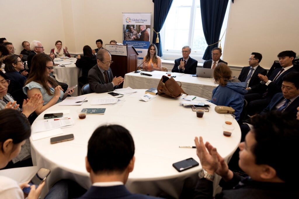 A group of people in business attire sit and clap around a large round table during the Capitol Policy Forum April 2026, held in a well-lit room as part of a formal meeting or conference.