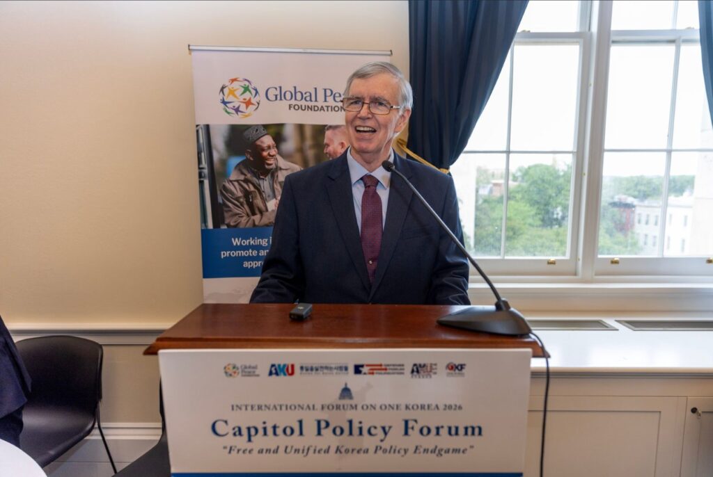An older man in a suit speaks at a podium during the Capitol Policy Forum, part of the International Forum on One Korea, with a Global Peace Foundation banner and a window in the background.