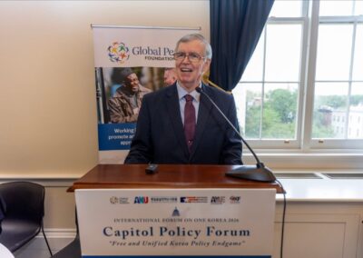 An older man in a suit speaks at a podium during the Capitol Policy Forum, part of the International Forum on One Korea, with a Global Peace Foundation banner and a window in the background.