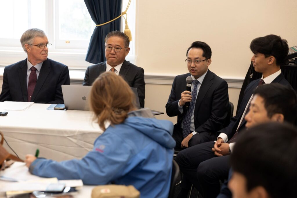Four men in business attire sit at a table, one speaking into a microphone—sharing Hyunseung Lee’s story of escape and freedom—as others listen intently and people in the foreground take notes.