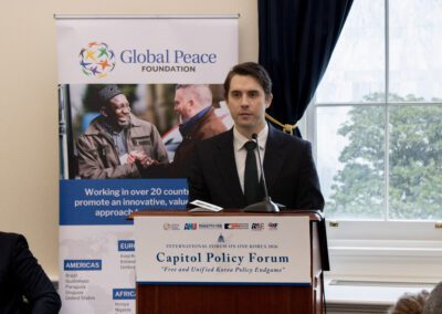 A man in a suit speaks at a podium during the Capitol Policy Forum, with an International Forum on One Korea banner from the Global Peace Foundation in the background.