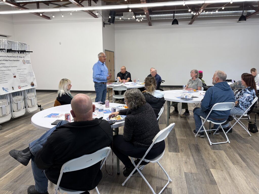 A man stands and speaks to a group of people seated around round tables in a bright, modern room with exposed ceilings, fostering cross community reconciliation.