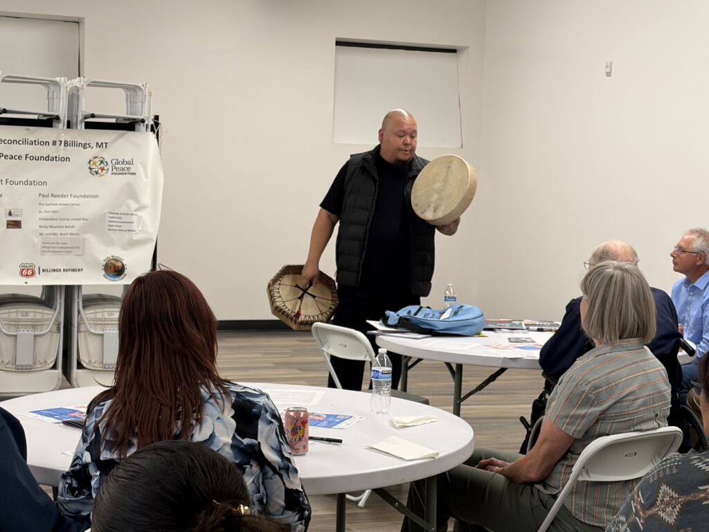 A man stands holding a hand drum and drumstick, sharing Indigenous voices and tradition with a seated group in a meeting room decorated with informational posters.