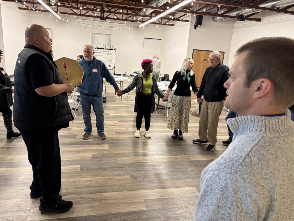 A group of people stand in a circle holding hands while a man with a drum addresses them, honoring Indigenous Voices, in a brightly lit room with exposed beams and tables in the background.