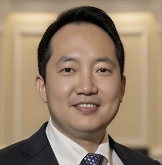 Hyun-Seung Lee, dressed in a suit and tie, smiles at the camera while standing indoors against a blurred, neutral background.
