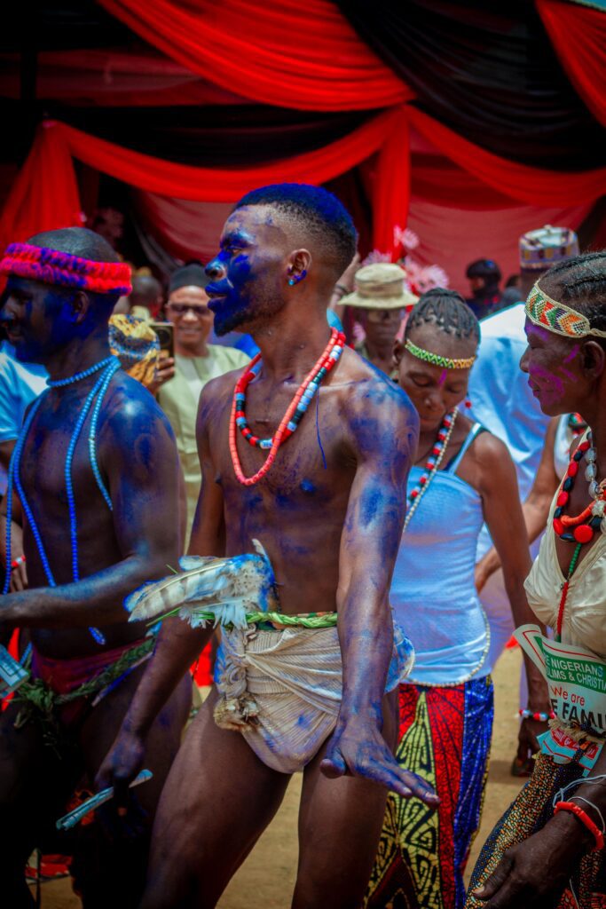 A group of people in traditional attire, some with blue body paint and beaded necklaces, participate in the Dung Nikyob Cultural Festival in Southern Kaduna, with red and black drapes in the background.