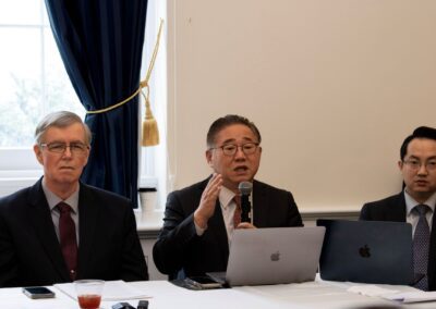 Three men in suits sit at a table in a conference room, with two laptops open; one man is speaking while the others listen attentively during the International Forum on One Korea.