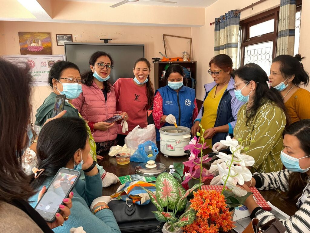 A group of women, some wearing masks, gather around a table with food and a rice cooker in a brightly lit room, discussing environmental stewardship and climate initiatives by GPF Nepal.
