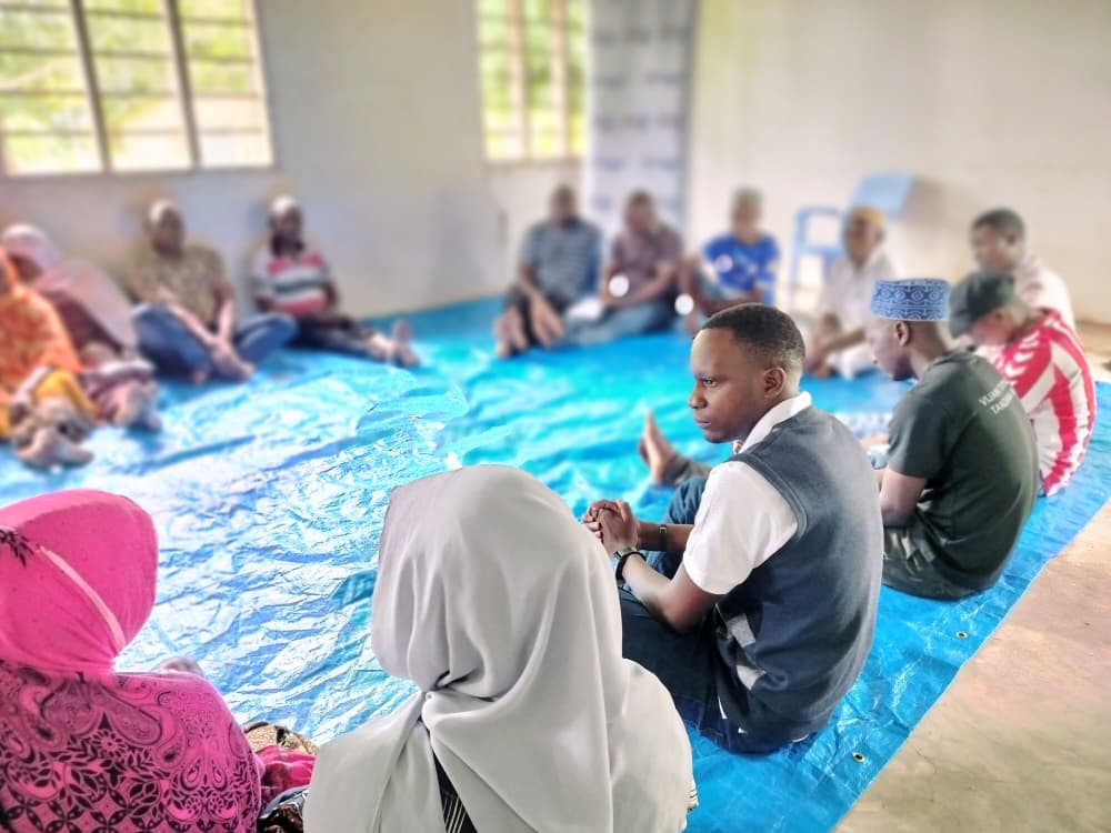 A group of people sit in a circle on a blue tarp inside a room, engaged in conversation about community development and transforming livelihoods, as part of a GPF Tanzania initiative.